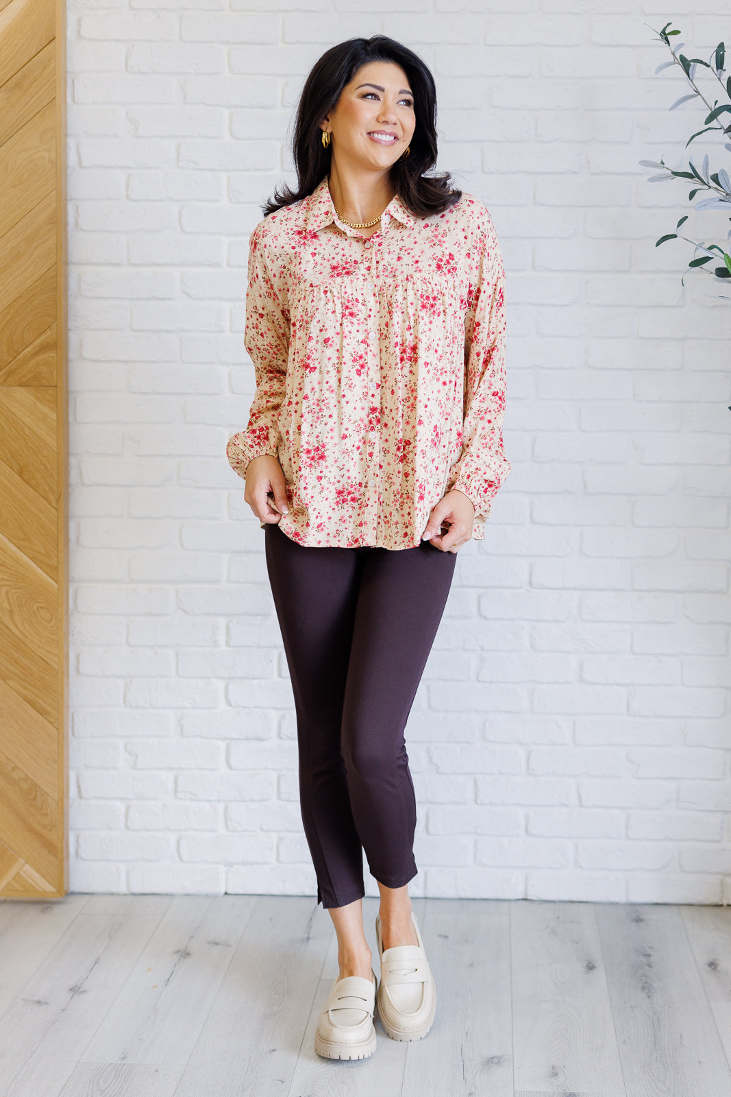 Woman wearing a floral blouse and dark pants against a white brick wall.