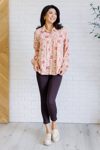 Woman wearing a floral blouse and dark pants against a white brick wall.