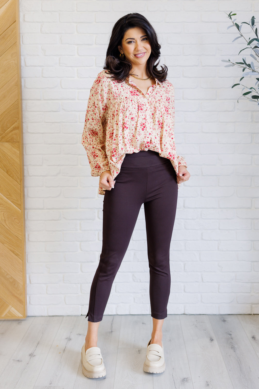 Woman wearing a floral blouse and dark pants standing against a white brick wall.
