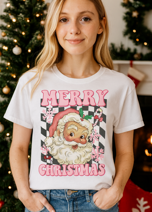 Person wearing a white t-shirt with a Christmas-themed design featuring Santa Claus, standing in front of a decorated Christmas tree.