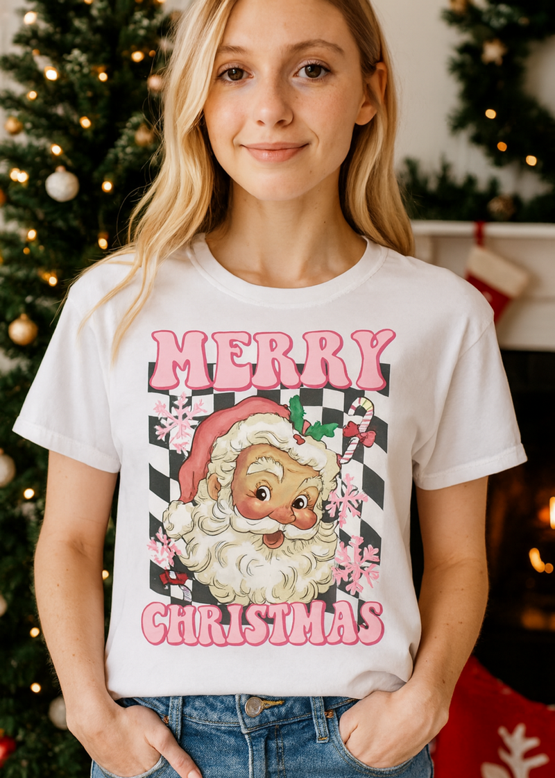 Person wearing a white t-shirt with a Christmas-themed design featuring Santa Claus, standing in front of a decorated Christmas tree.