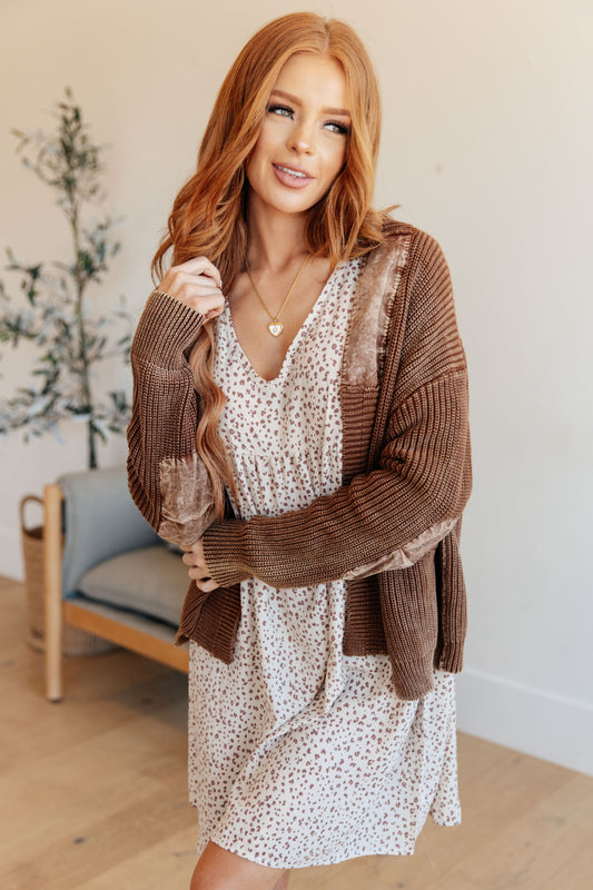 Woman wearing a brown cardigan over a white dress with a floral pattern, standing indoors.