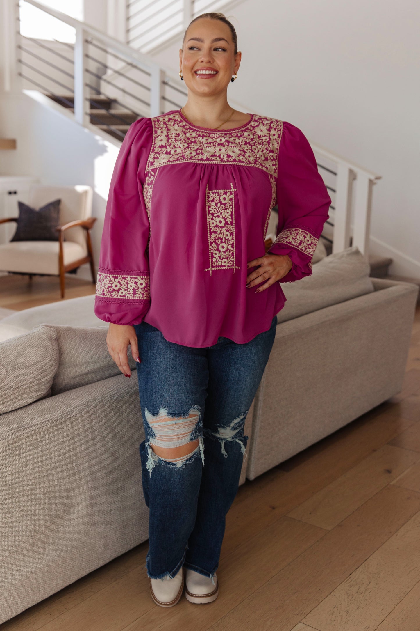 Woman wearing a pink blouse with floral patterns and ripped jeans in a living room.