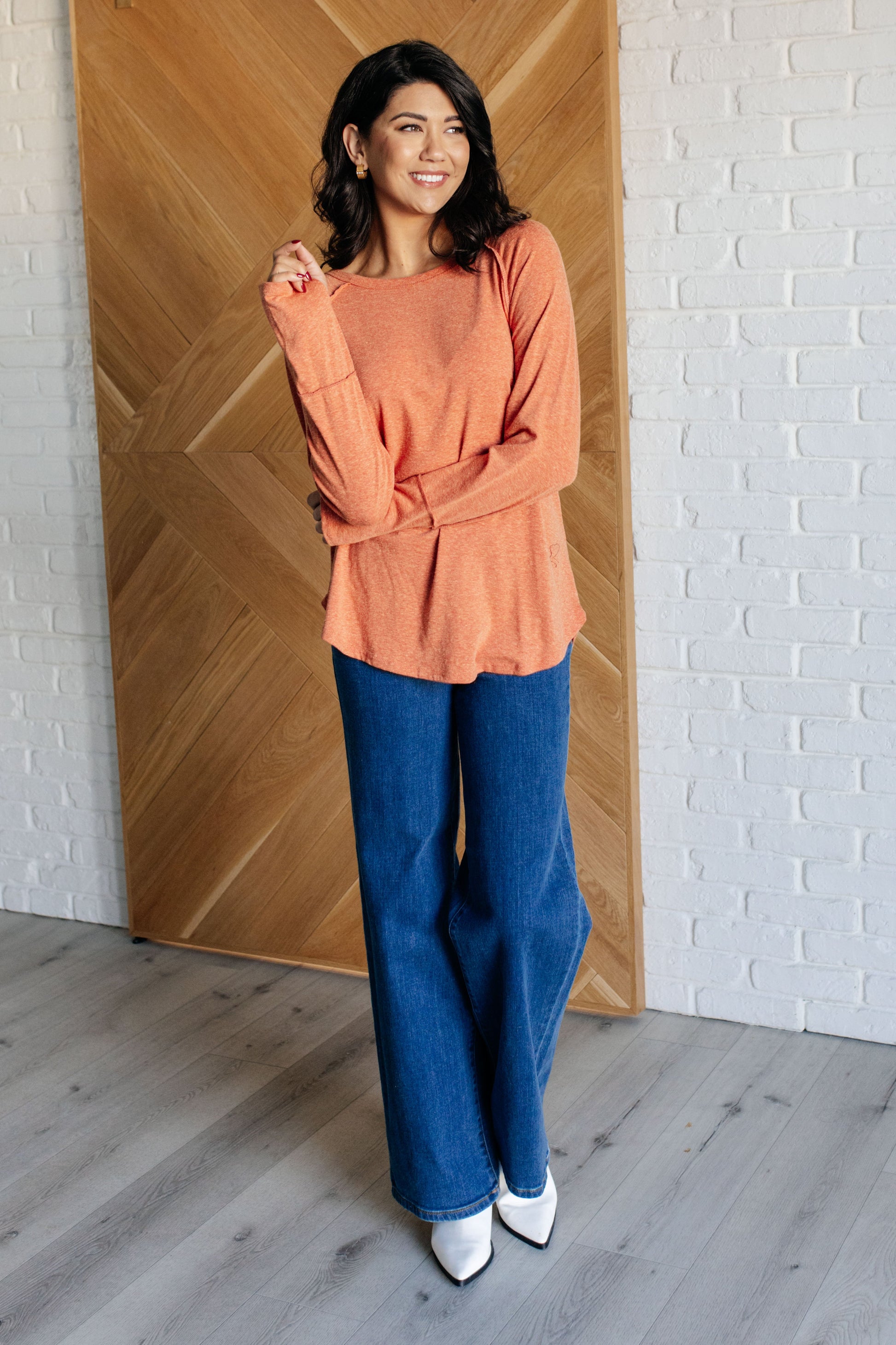 Woman wearing an orange top and blue jeans standing in a room with wooden paneling and white brick wall.