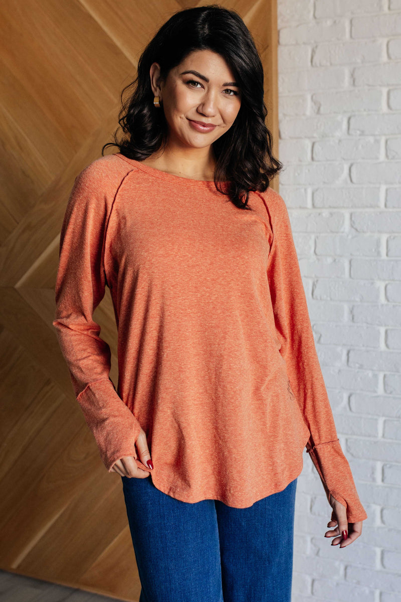 Woman wearing an orange long-sleeve top and blue pants against a white brick wall.