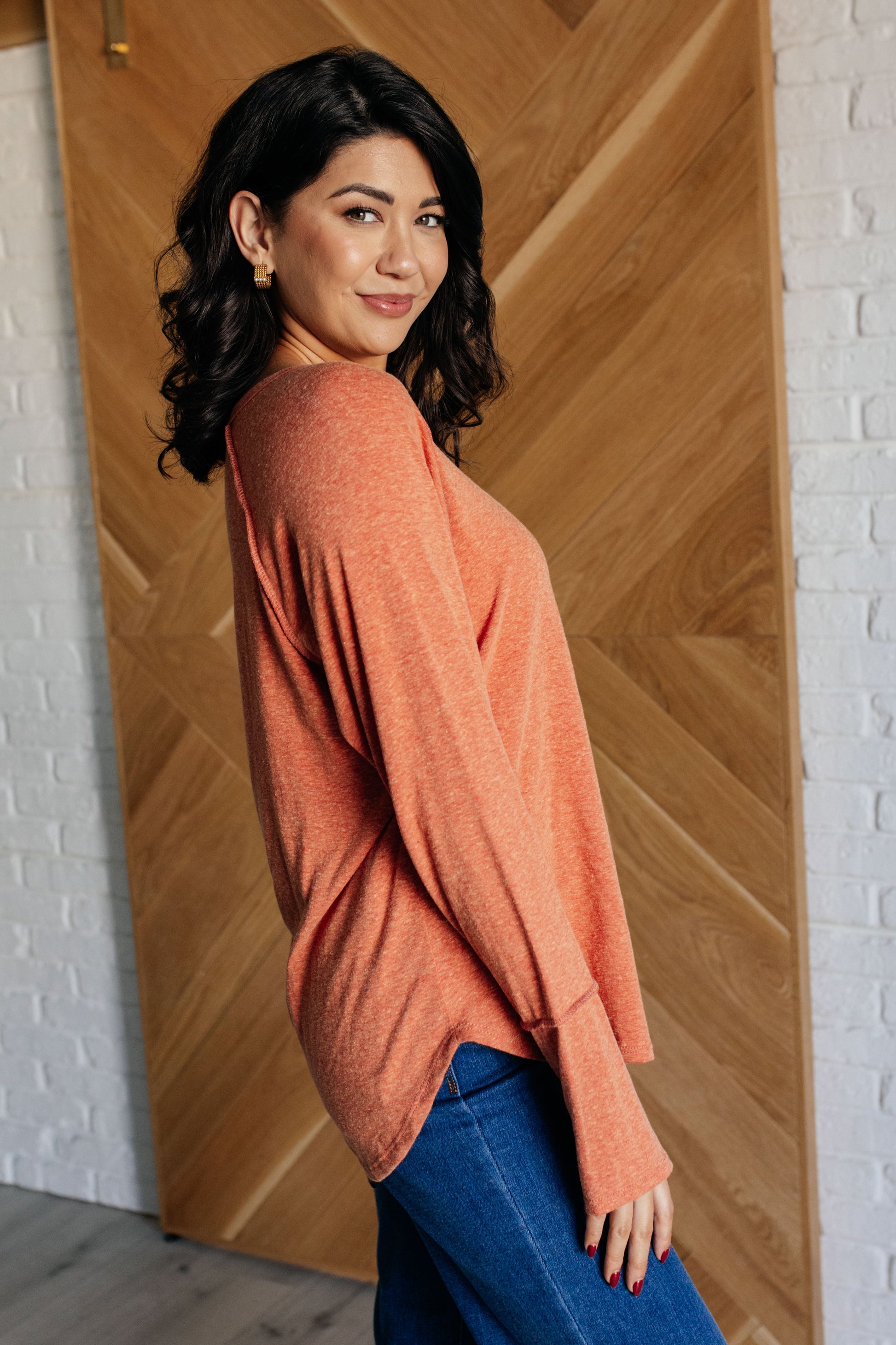 Woman wearing an orange sweater and blue jeans standing against a wooden wall.