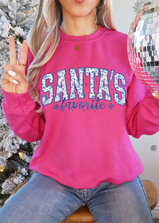 Person wearing a pink shirt with 'Santa's Favorite' text, holding a disco ball in front of a decorated Christmas tree.