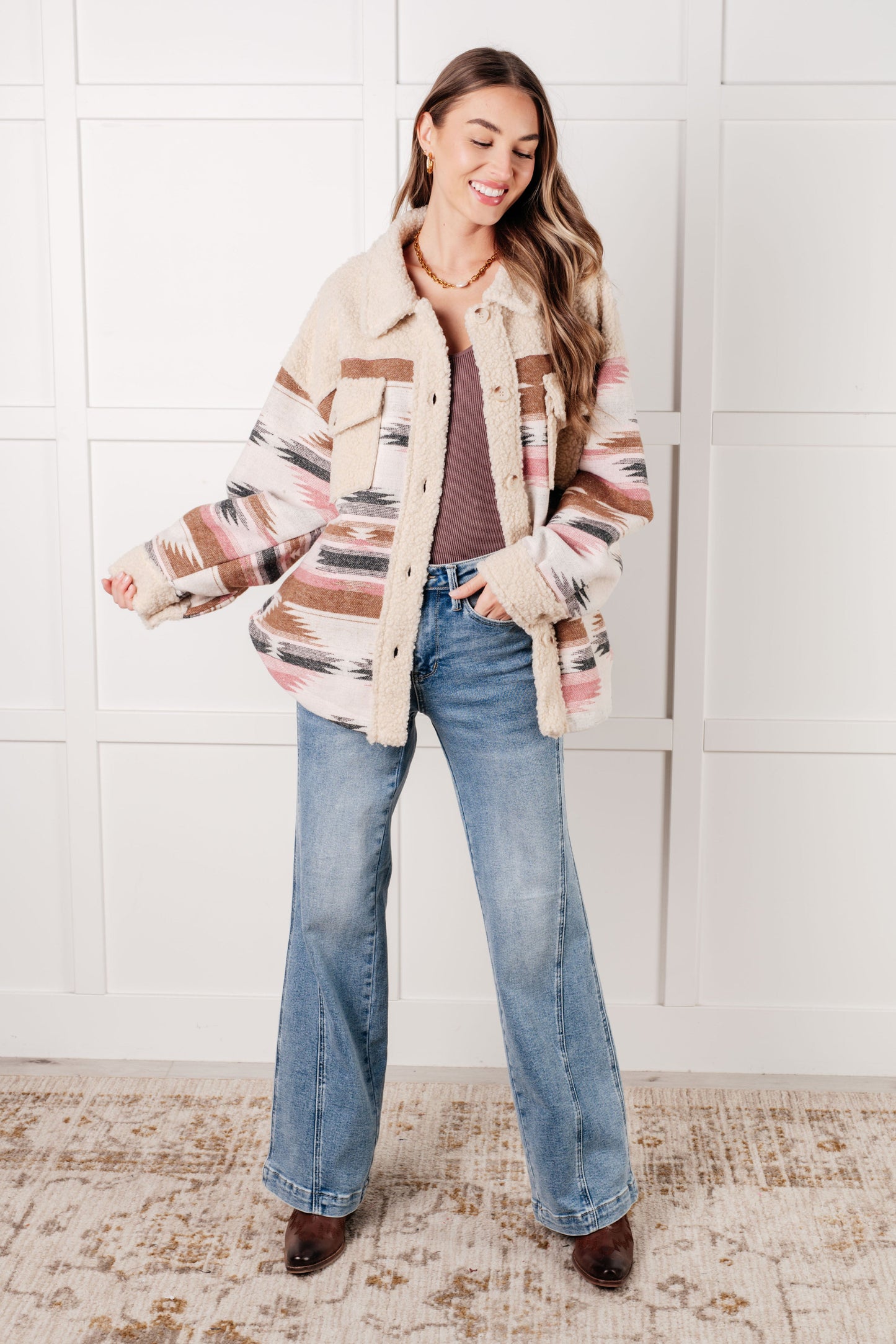 Woman wearing a patterned jacket and jeans standing against a white tiled wall.