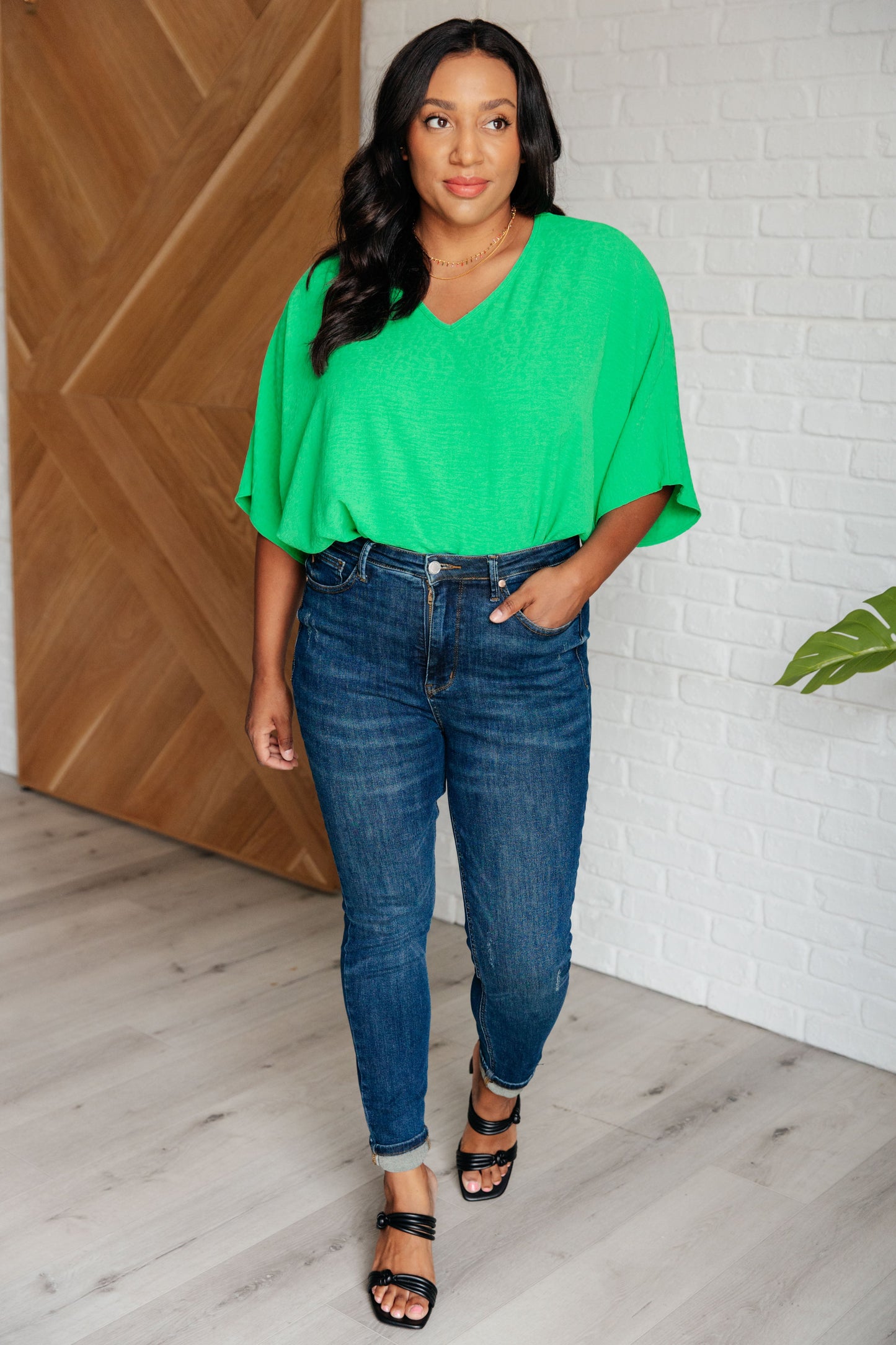 Woman wearing a green top and blue jeans standing in a room with wooden and white brick walls.