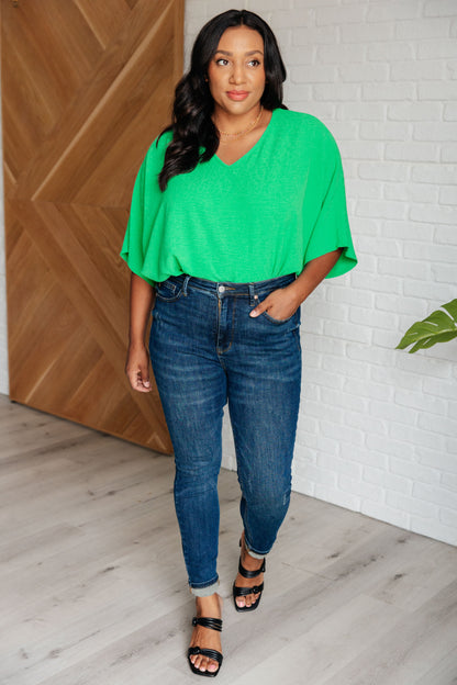 Woman wearing a green top and blue jeans standing in a room with wooden and white brick walls.