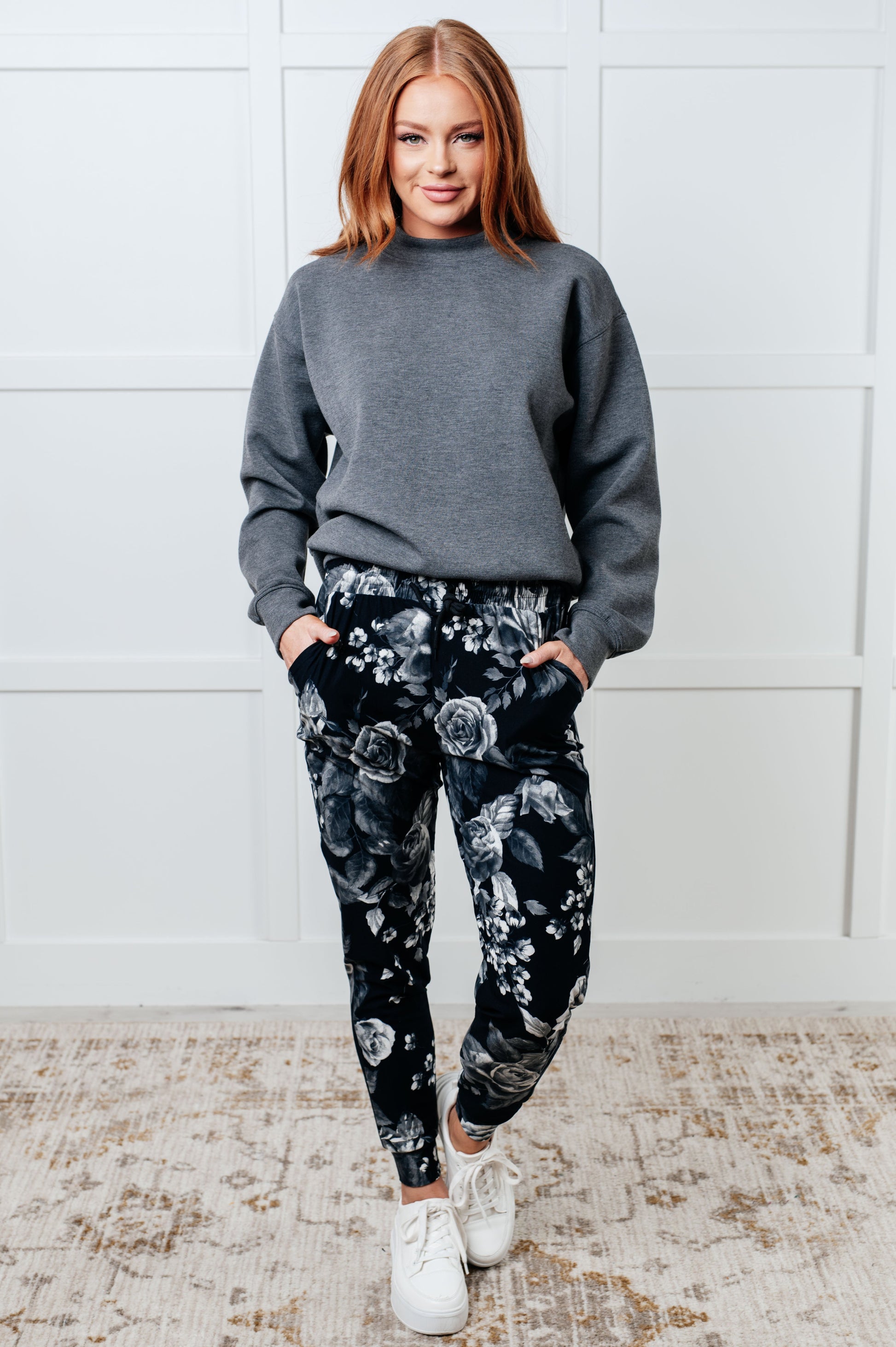 Woman wearing a gray sweater and black floral pants against a white wall.