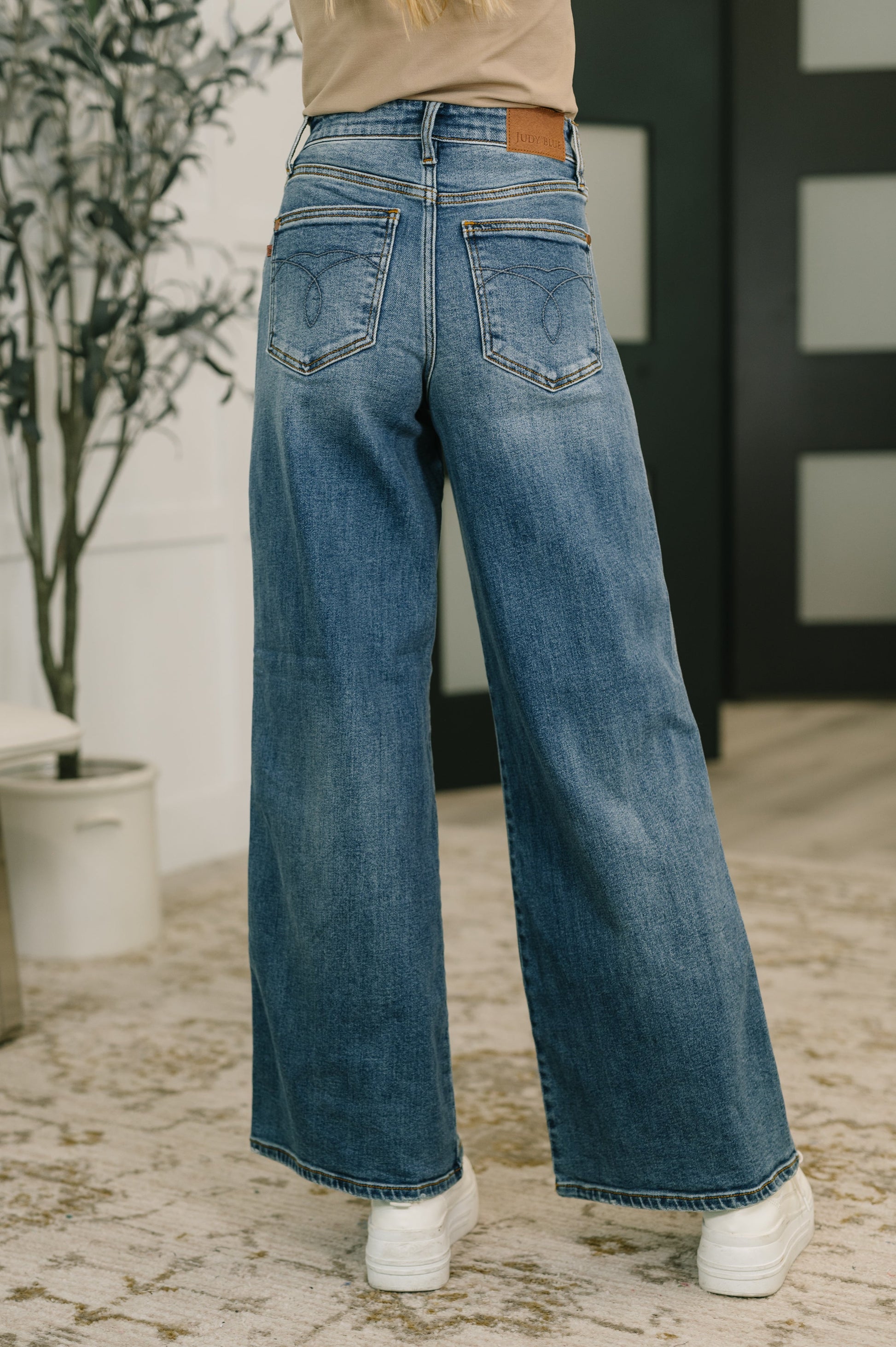 Person wearing blue jeans standing indoors with a plant and wall in the background