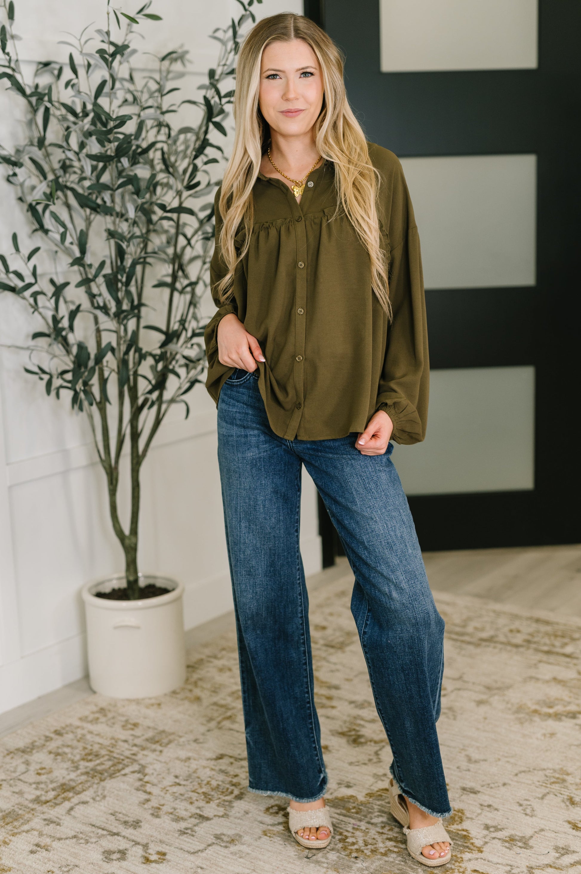 Woman wearing an olive green blouse and blue jeans standing indoors with a plant in the background.