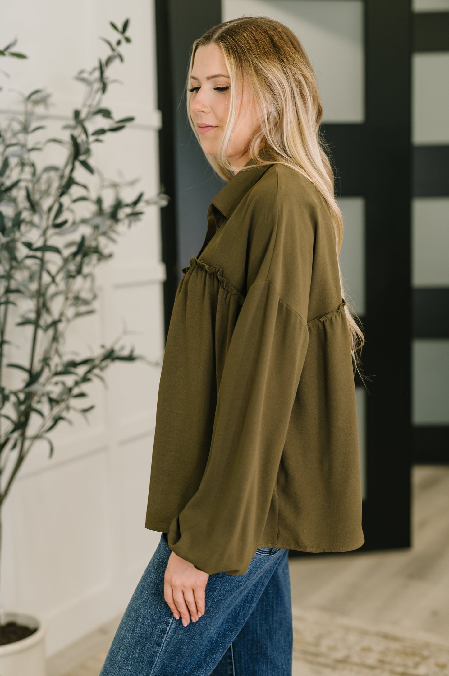 Woman wearing an olive green jacket and blue jeans standing indoors.