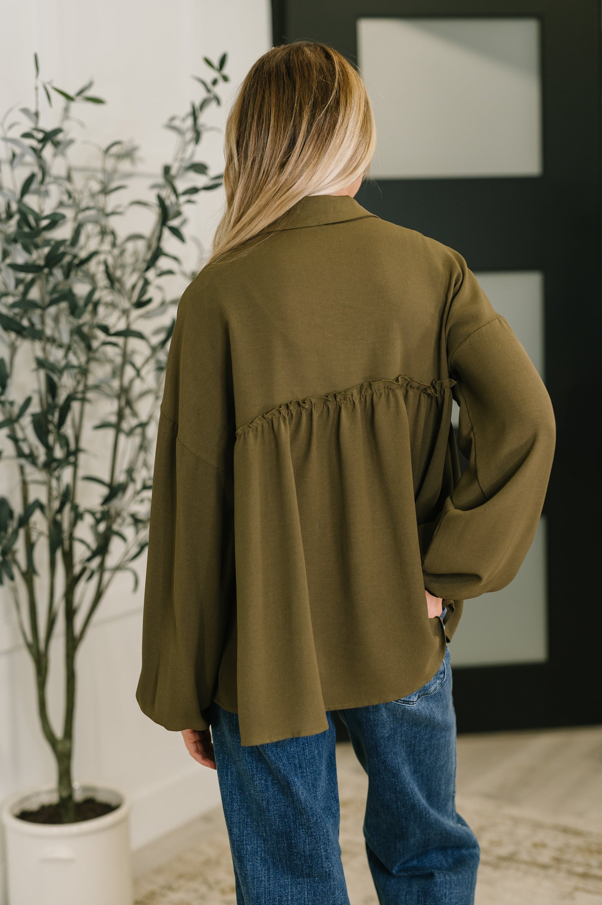 Person wearing an olive green jacket and blue jeans standing indoors with a plant and framed picture in the background.