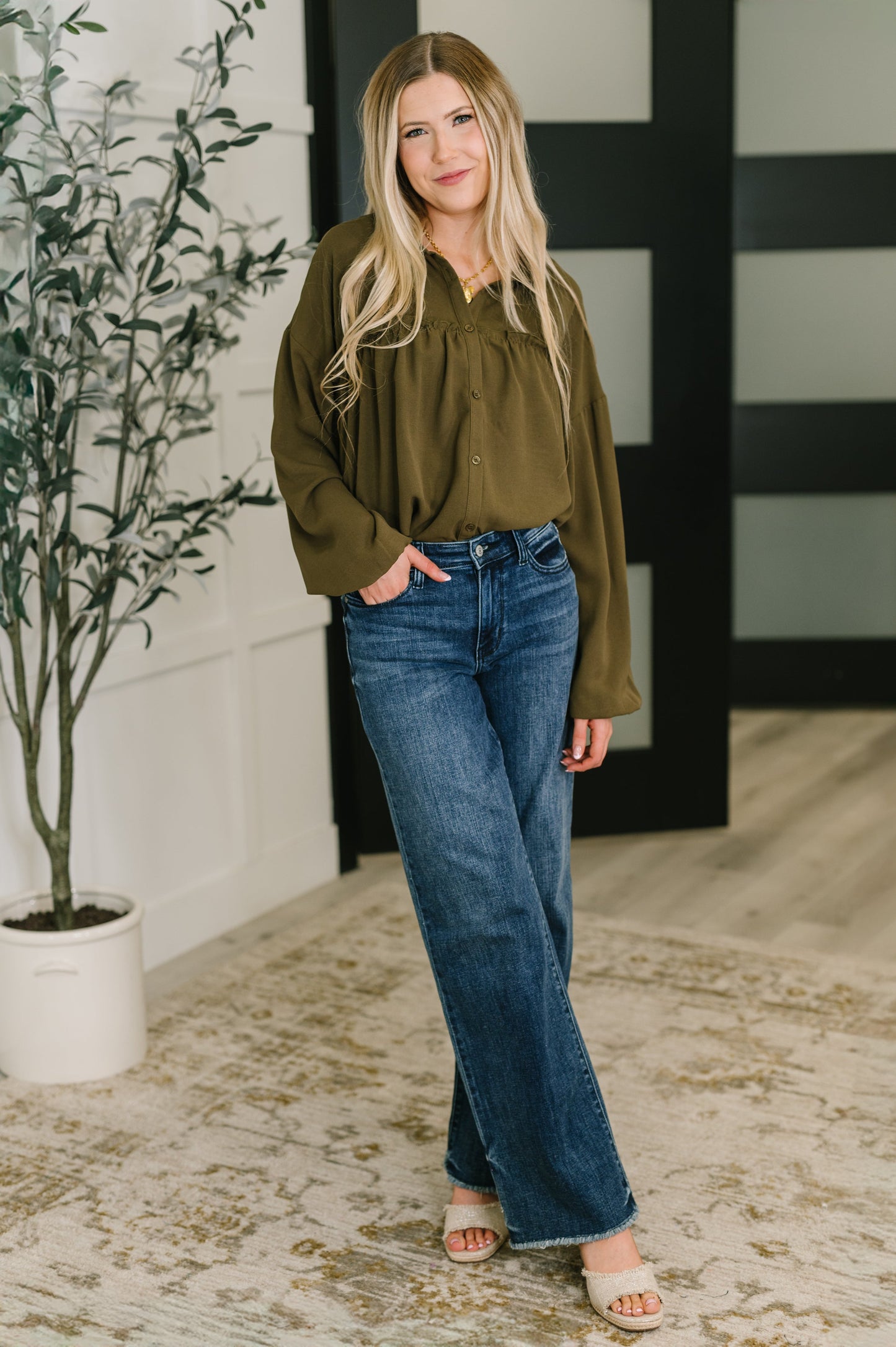 Woman wearing a green blouse and blue jeans standing indoors with a plant and decorative wall in the background.