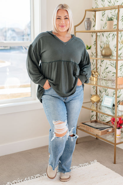 Woman wearing a green sweater and ripped jeans in a room with a bookshelf and window.