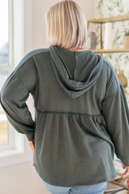Person wearing a green hooded jacket in a room with a window and shelves.