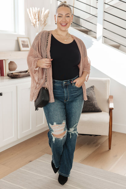 Woman in a casual outfit with a pink cardigan, black top, and ripped jeans standing in a modern living room.