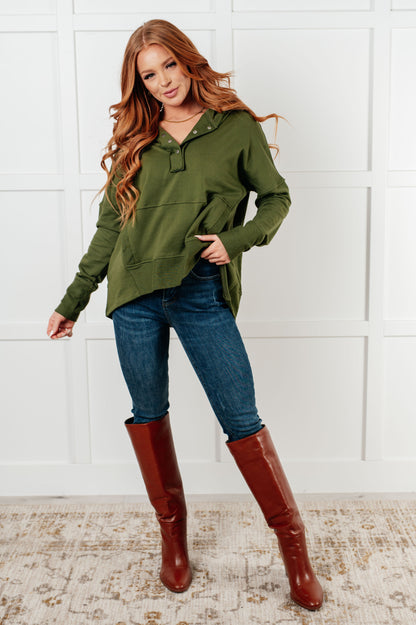 Woman wearing an olive green hoodie, blue jeans, and brown knee-high boots against a white brick wall.