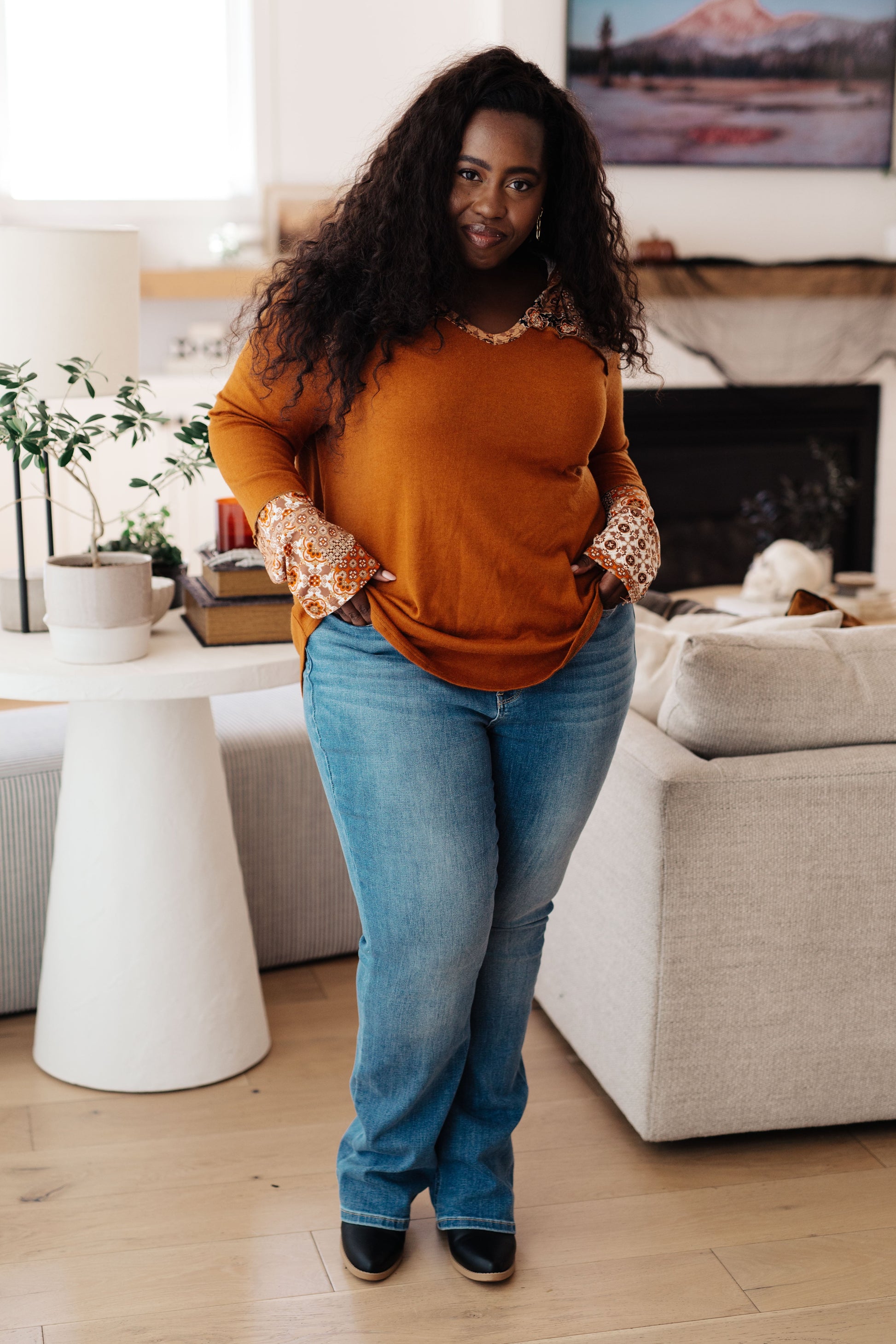 Woman standing in a living room wearing an orange sweater and blue jeans.
