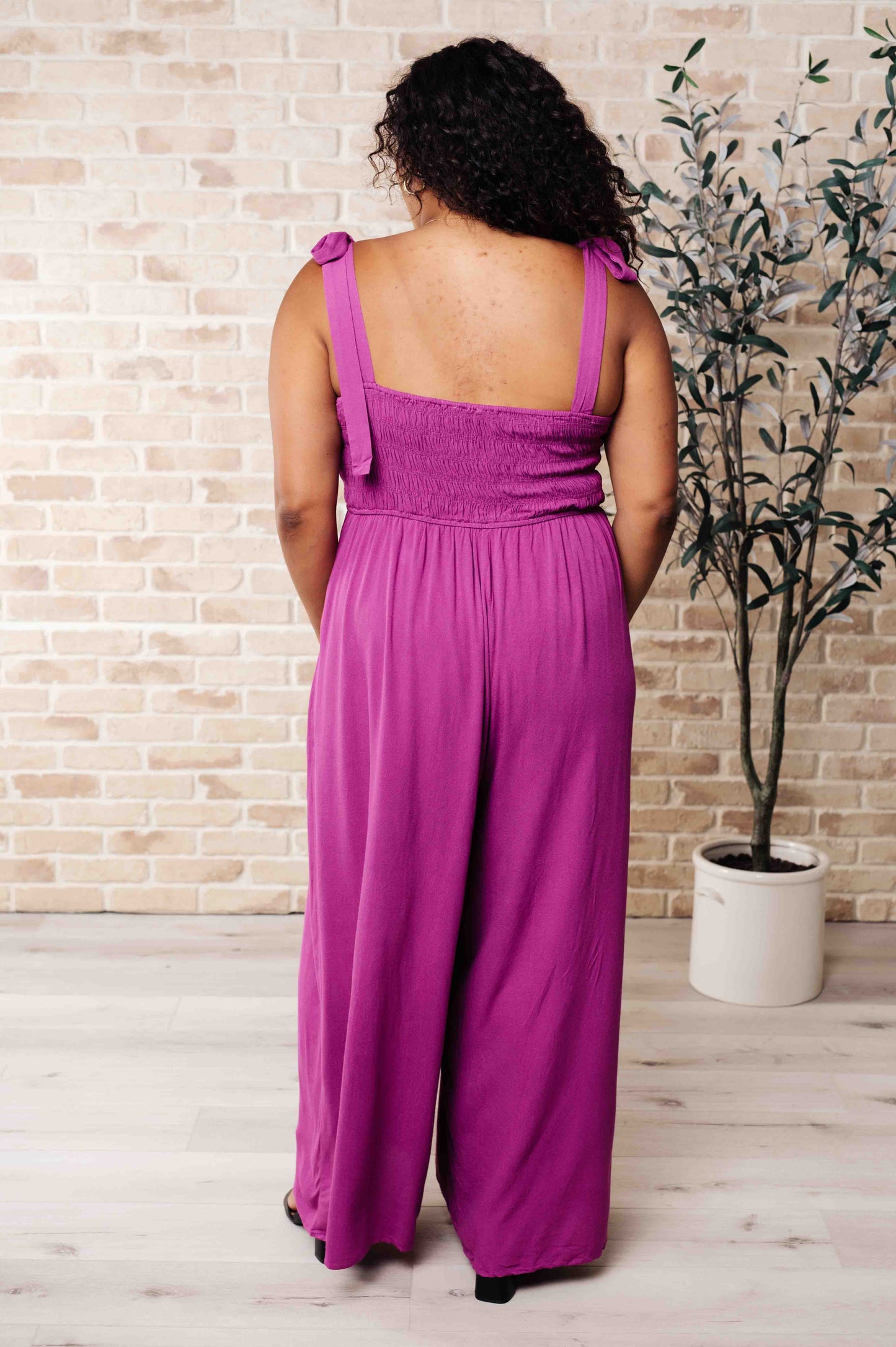 Person wearing a purple jumpsuit standing in front of a brick wall with a plant.