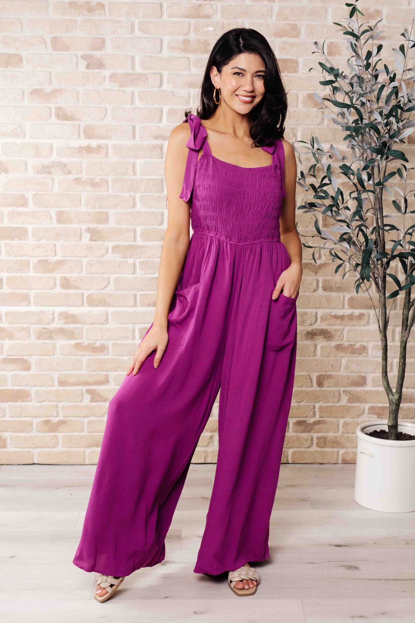 Woman wearing a purple jumpsuit standing in front of a brick wall with a plant.