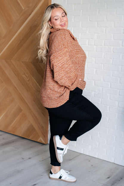 Woman wearing a brown sweater and black pants posing against a white brick wall.