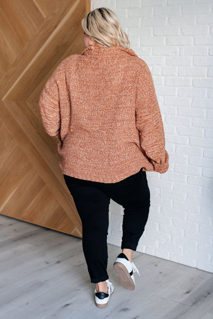 Person wearing a brown textured sweater and black pants standing against a white brick wall.