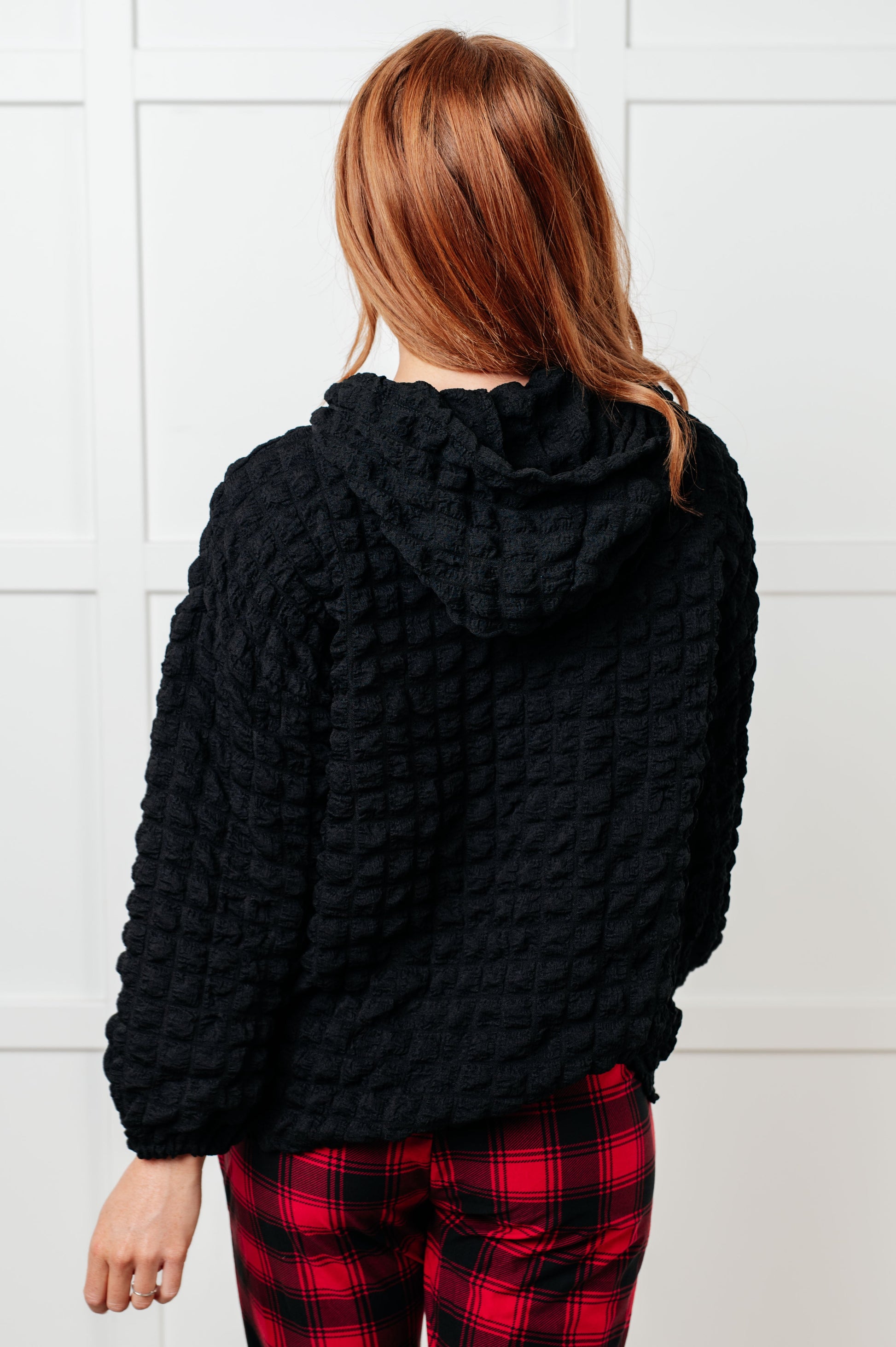 Person wearing a black textured sweater and red plaid pants against a white tiled wall.