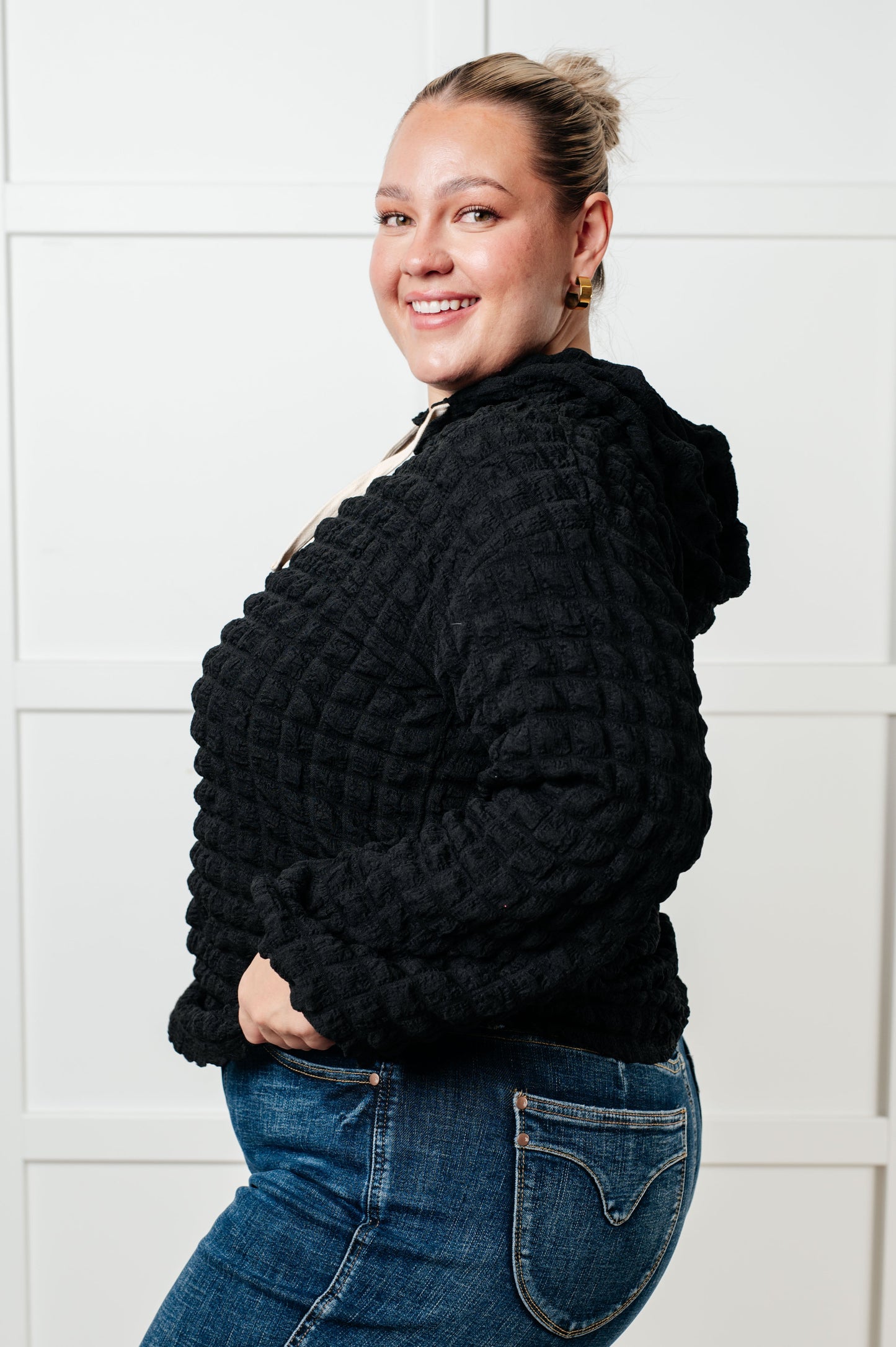 Woman wearing a black textured sweater and blue jeans against a white tiled wall.