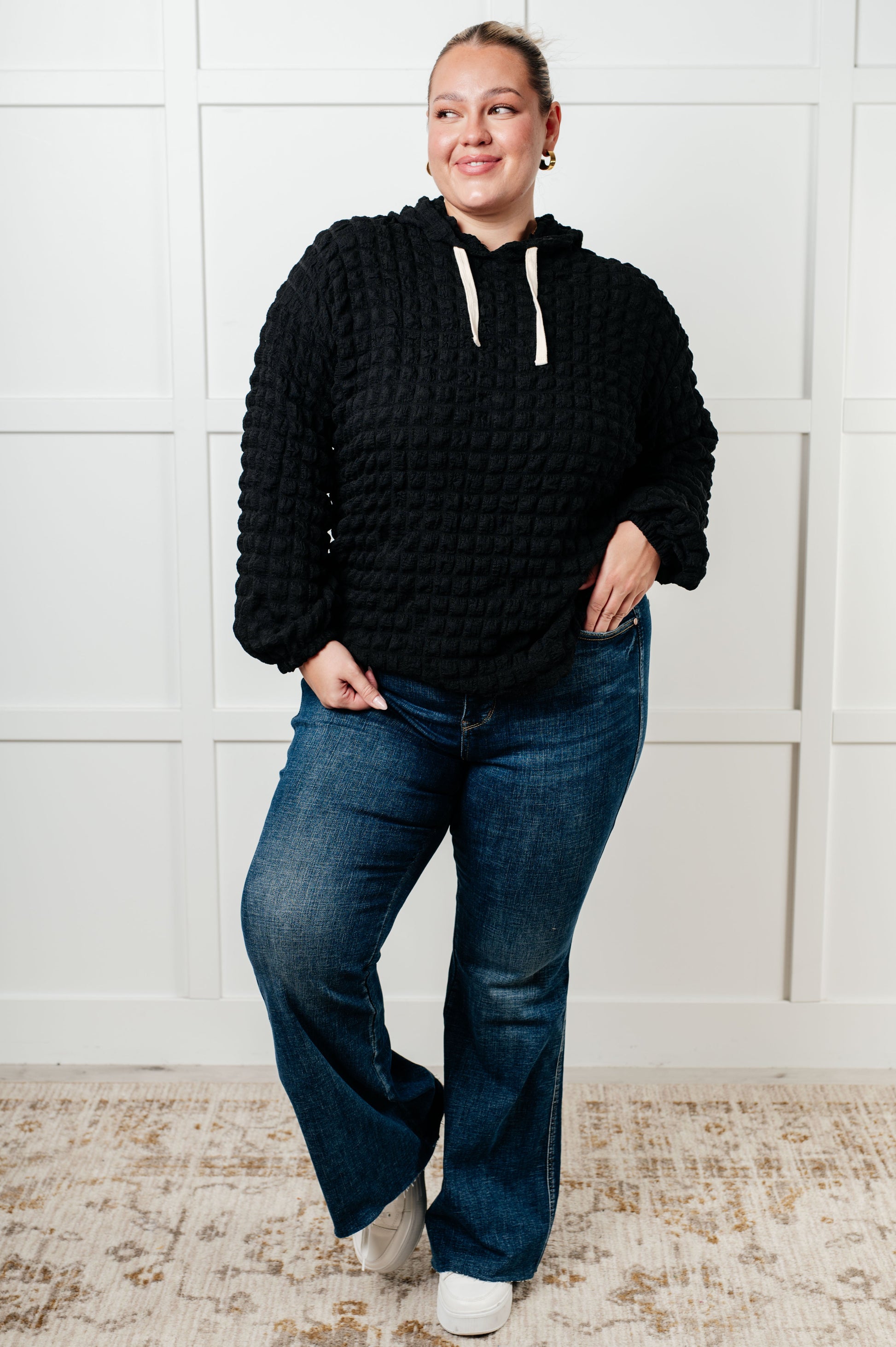 Person wearing a black textured hoodie and blue jeans standing against a white paneled wall.