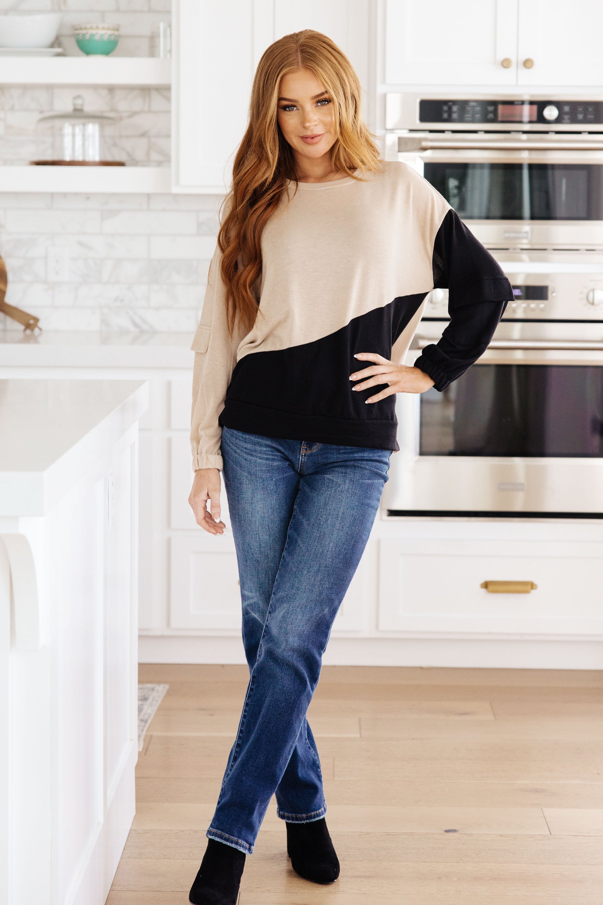 Woman wearing a beige and black sweater with blue jeans in a kitchen.