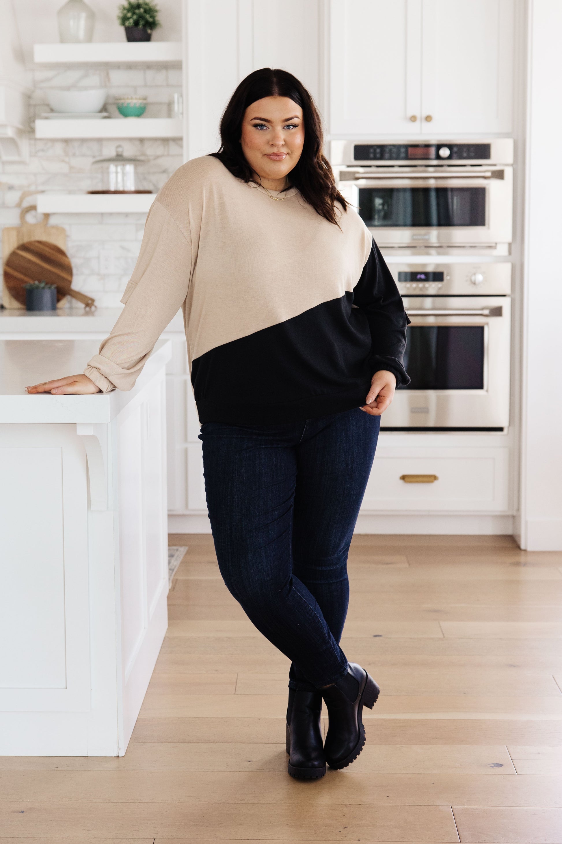 Woman standing in a kitchen wearing a beige and black sweater with dark jeans.
