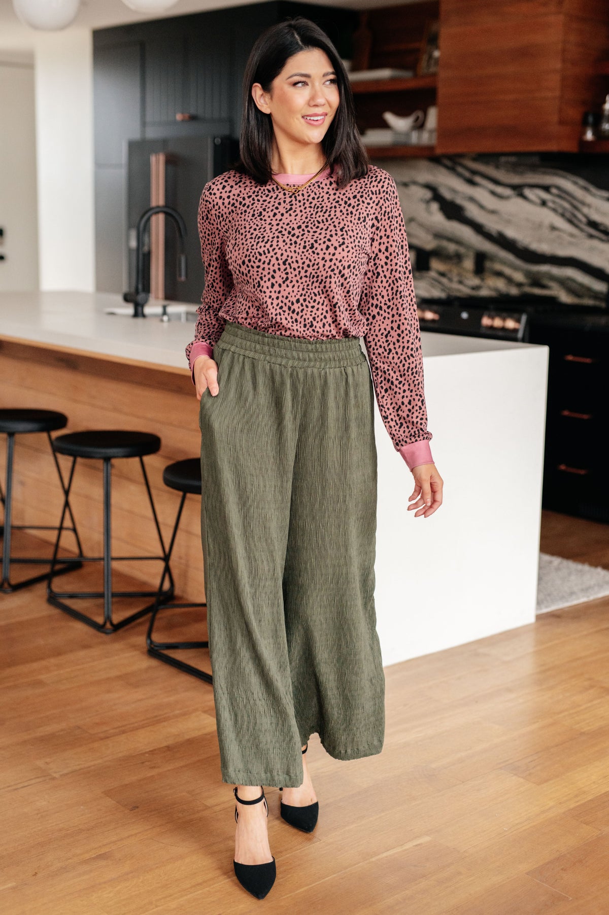 Woman in a kitchen wearing a patterned top and green pants.