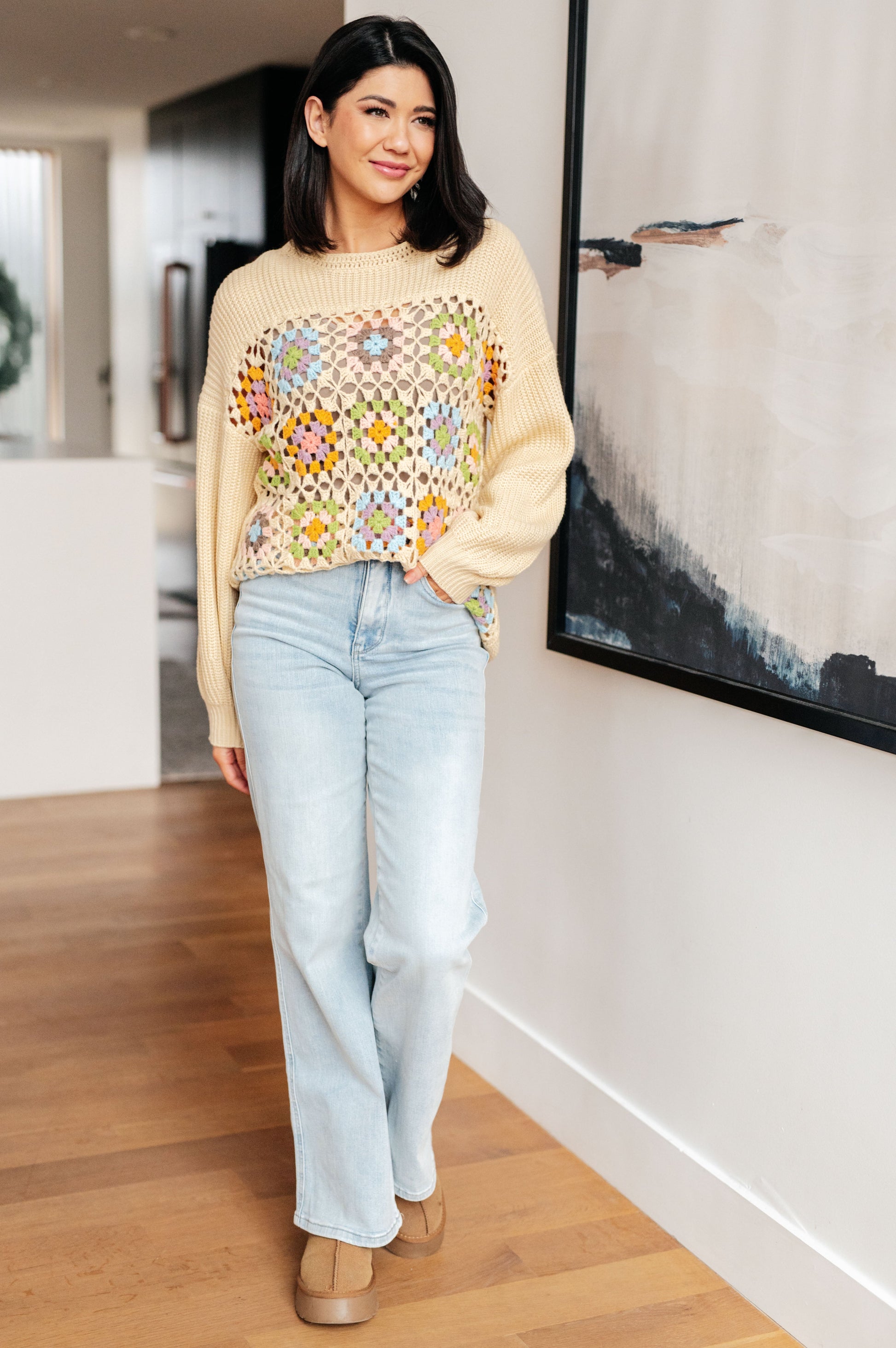 Woman wearing a colorful sweater and light blue jeans standing in a room with wooden flooring and a wall art piece.