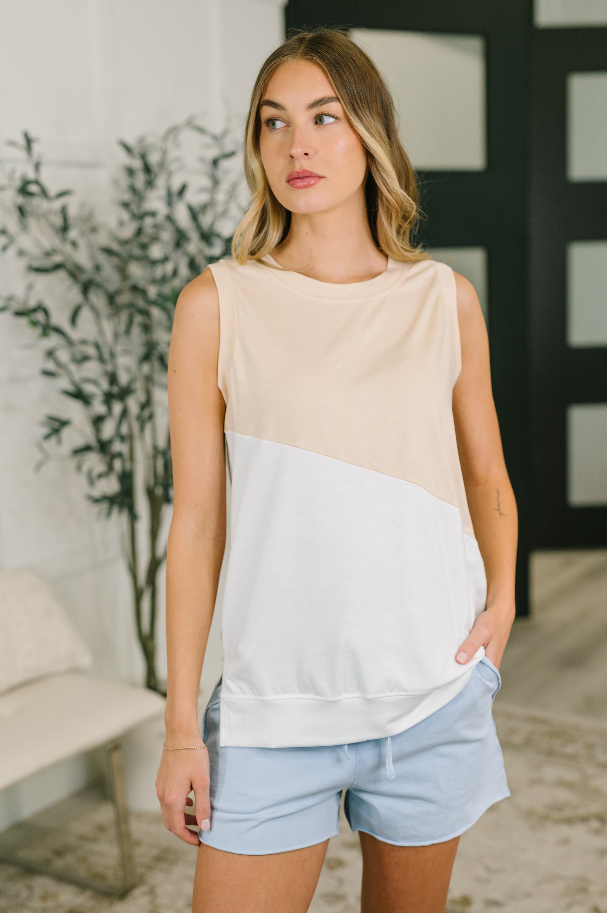 Woman wearing a beige and light gray sleeveless top with light blue shorts indoors.