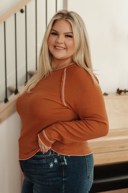 Woman wearing a rust-colored sweater and blue jeans standing indoors.