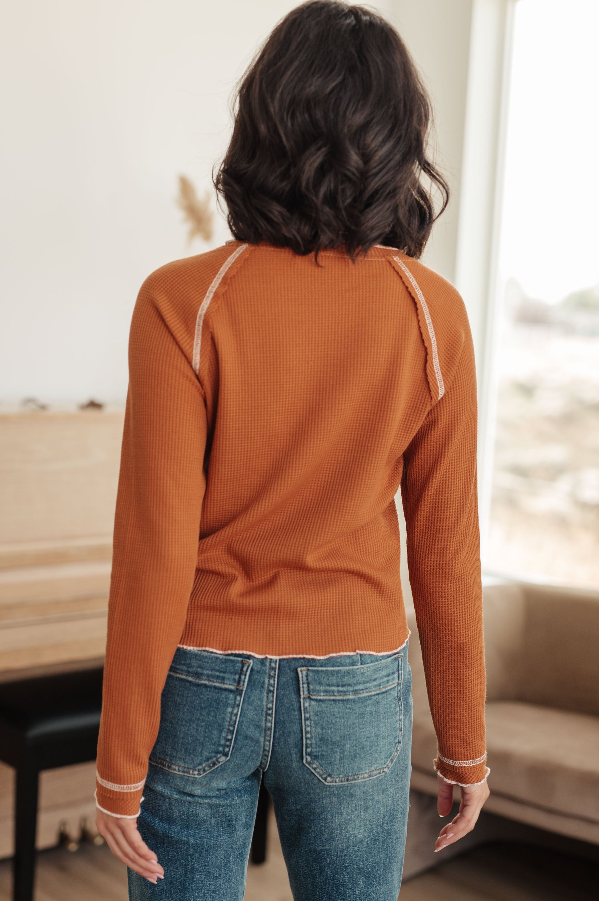 Person wearing an orange long-sleeve top and blue jeans in a room with a piano.