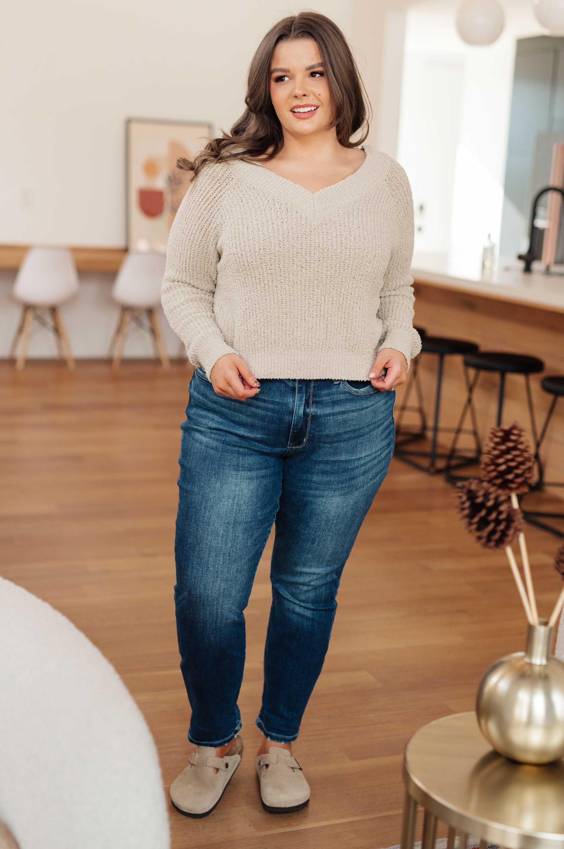 Woman wearing a beige sweater and blue jeans standing in a room with wooden flooring and decor.