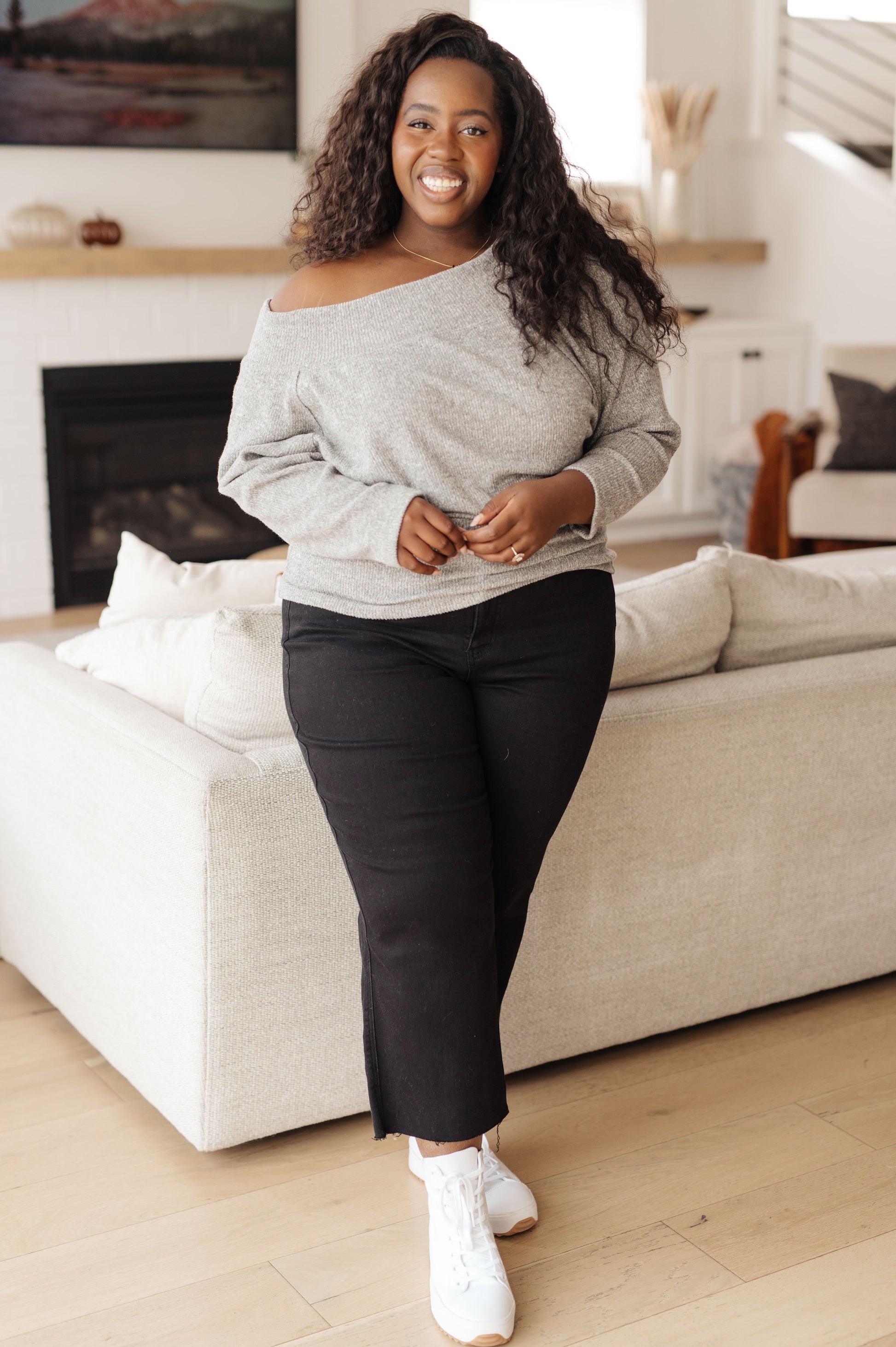 Woman in a gray off-shoulder top and black pants standing in a living room.
