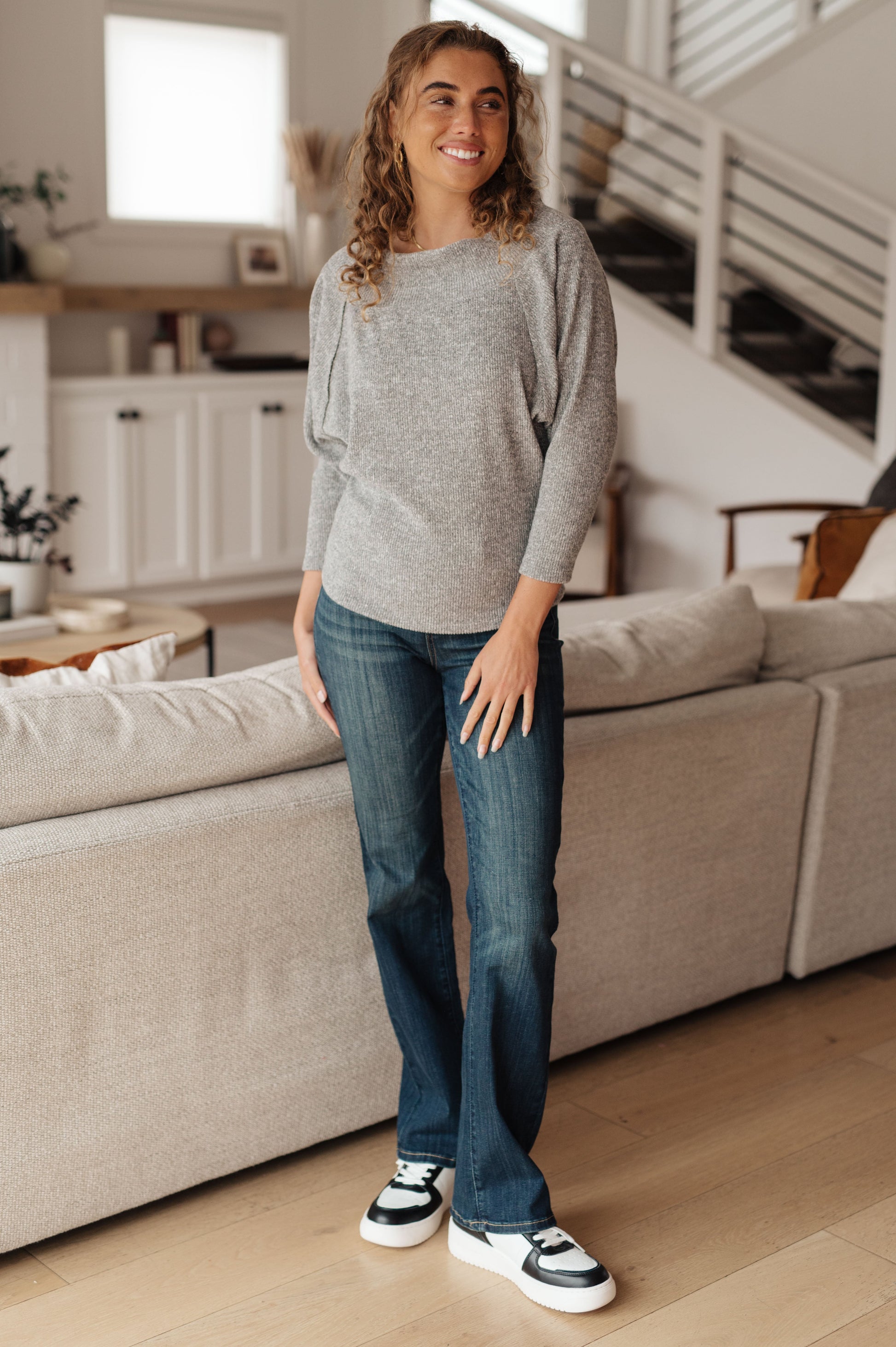 Woman standing in a living room wearing a gray sweater and blue jeans.