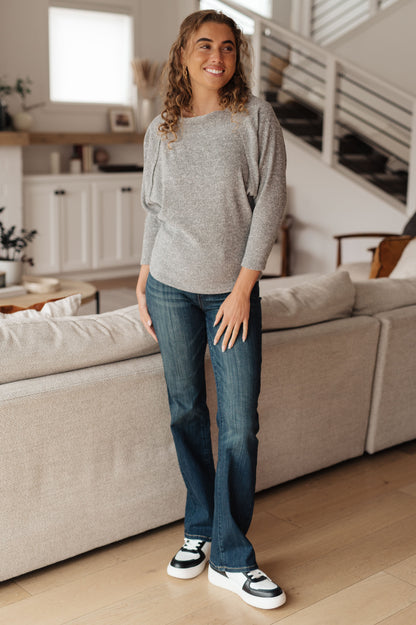 Woman standing in a living room wearing a gray sweater and blue jeans.