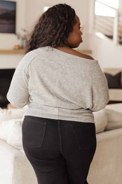 Person wearing a gray off-shoulder top and dark pants in a living room setting.
