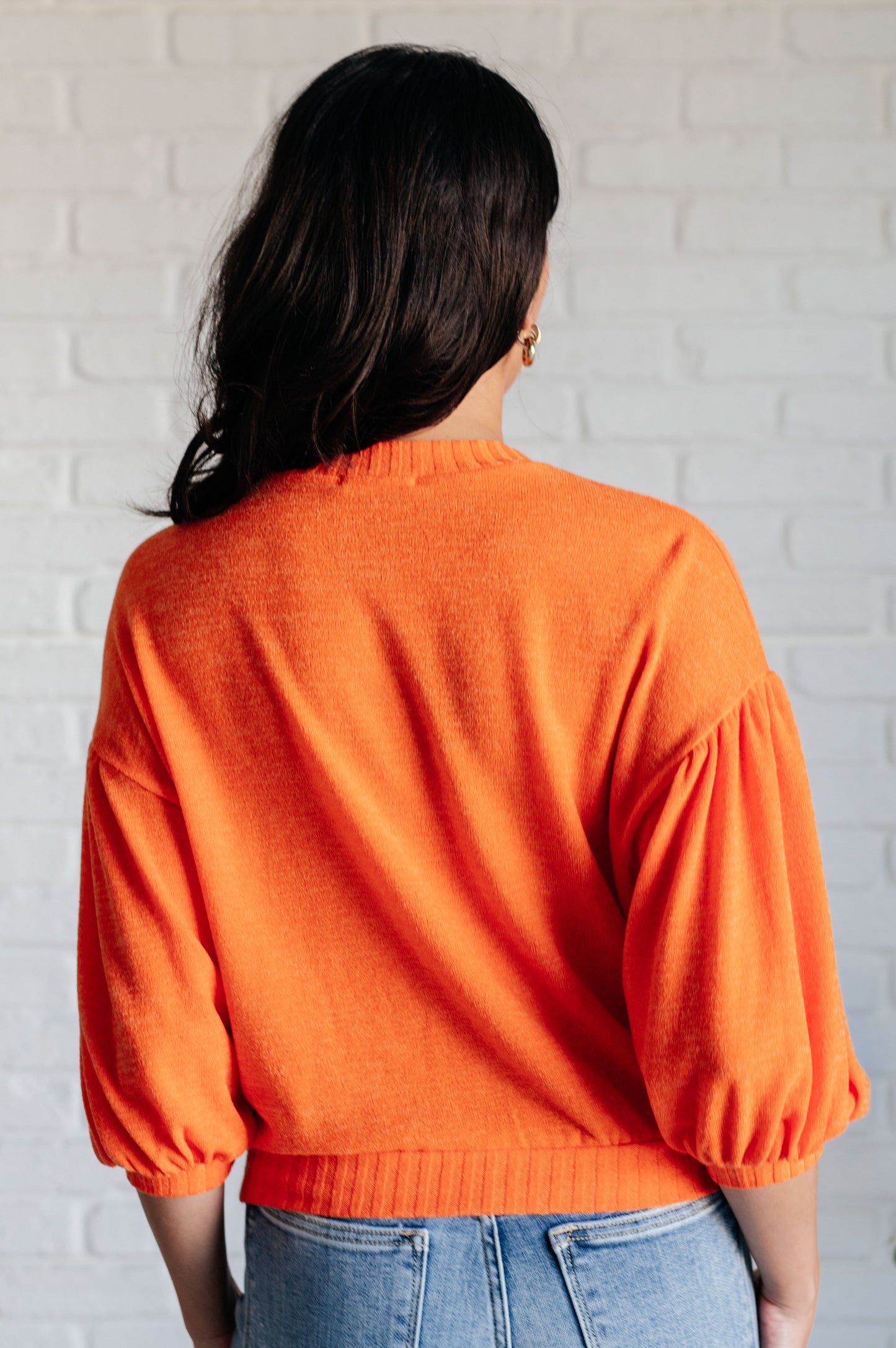 Person wearing an orange sweater with balloon sleeves against a white brick wall.