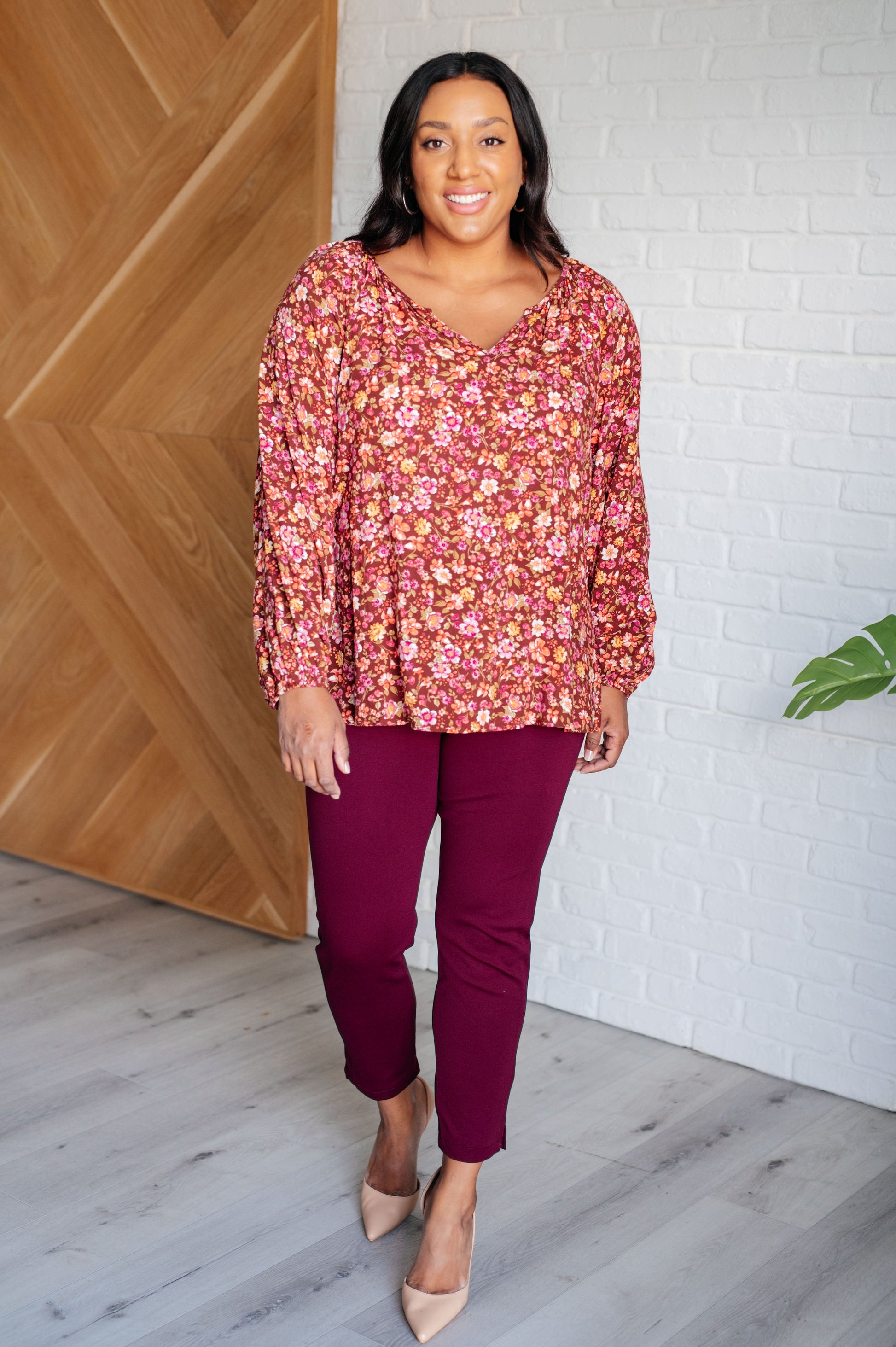 Woman wearing a floral blouse and purple pants standing against a wooden and white brick wall.