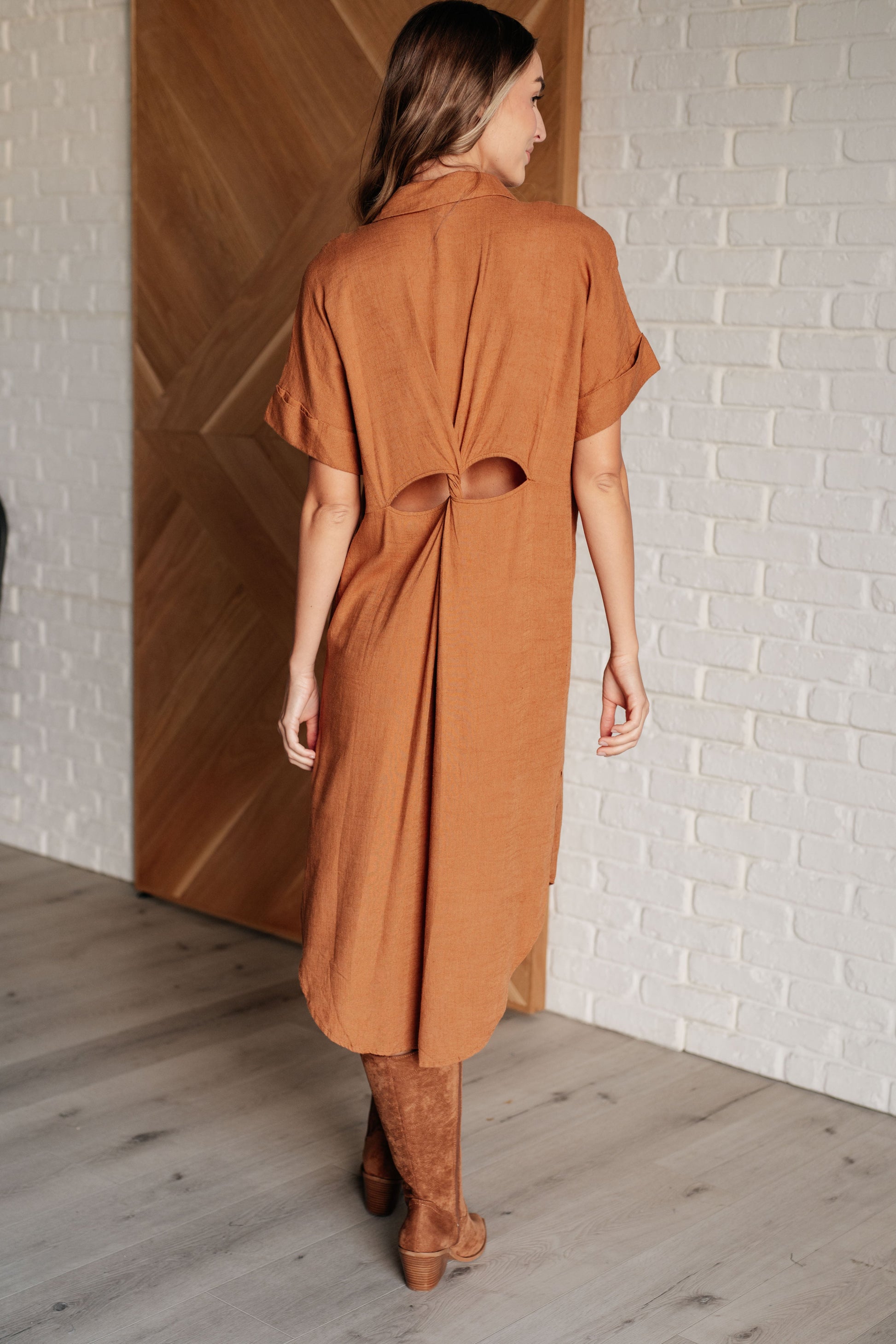Woman wearing a brown dress standing in a room with wooden flooring and a white brick wall.