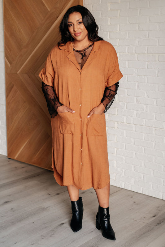 Woman wearing a mustard yellow dress with black sleeves and boots against a white brick wall.