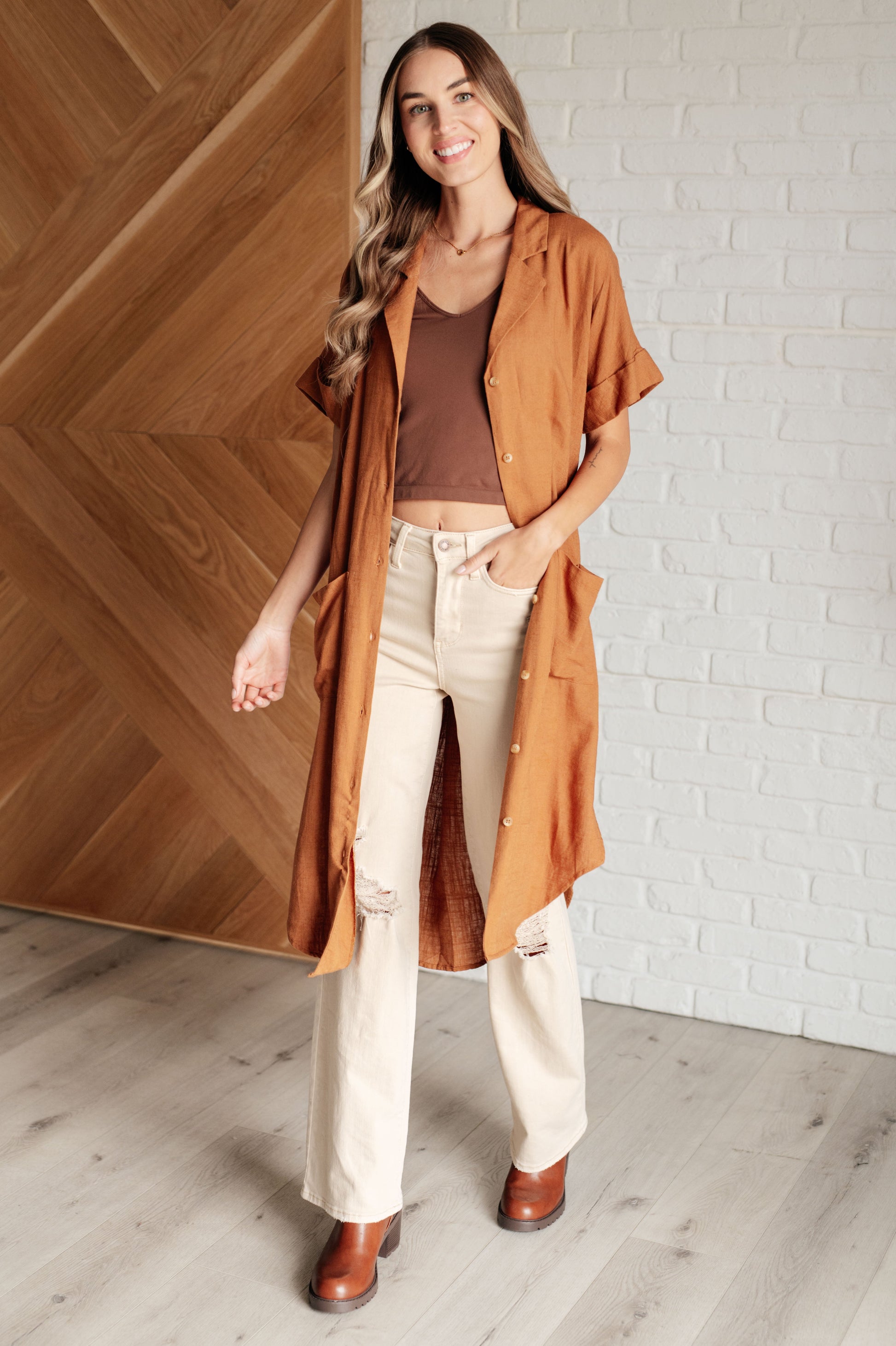 Woman wearing a long brown coat over a brown top and white pants, standing against a wooden wall.