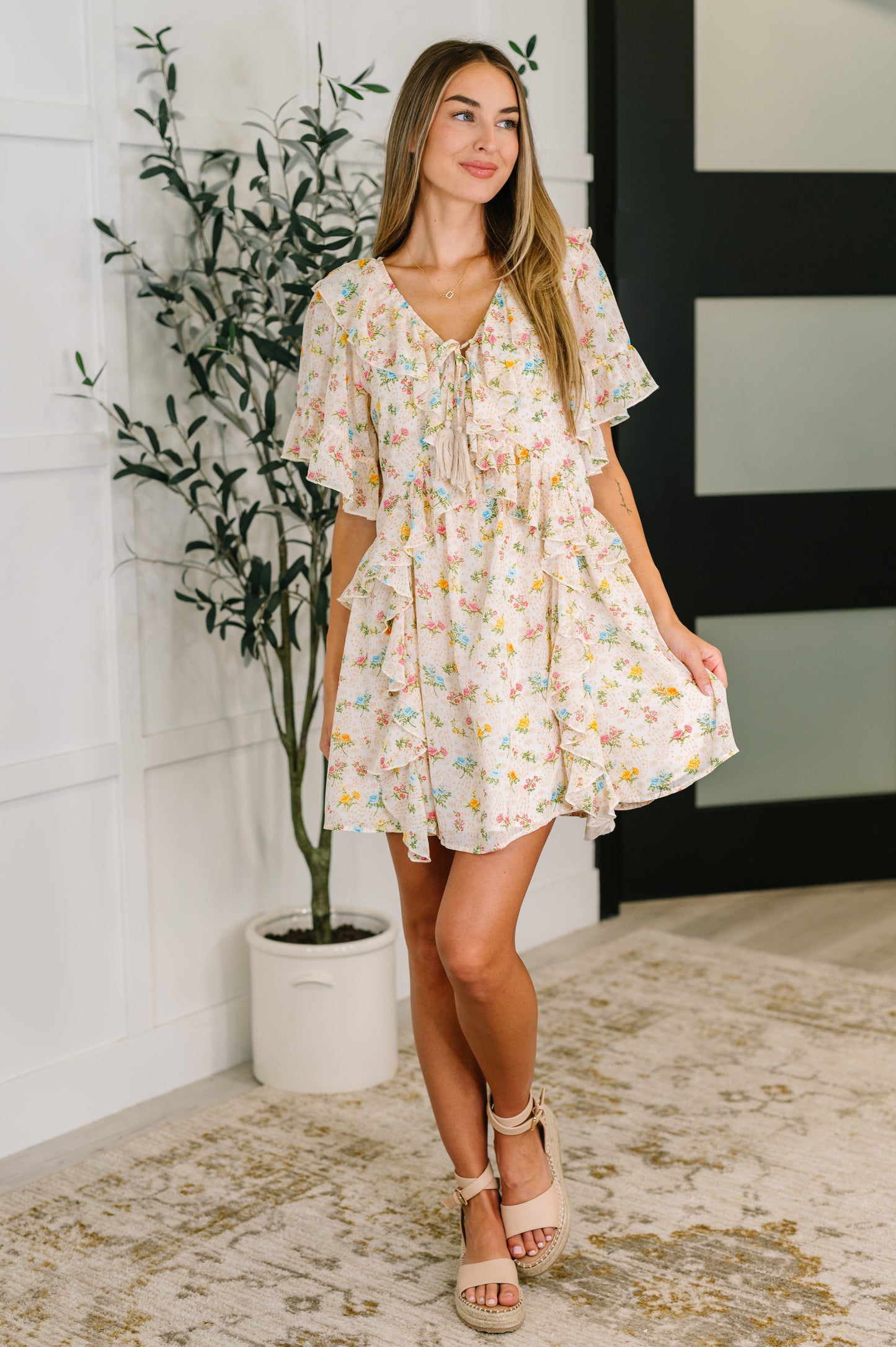 Woman wearing a floral dress standing indoors with a plant and wall in the background
